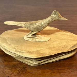 May include: A carved wooden sculpture of a roadrunner bird perched on a layered, square-shaped base. The bird is light brown with detailed feathering, and the base has a textured, natural appearance. The sculpture is set against a dark wood surface.