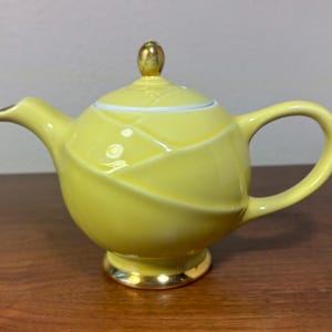 May include: A yellow ceramic teapot with a gold-colored handle, spout, and base. The lid is topped with a gold knob. The teapot has a unique design with curved, overlapping sections. The teapot is sitting on a wooden surface.