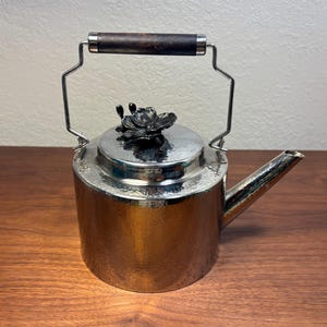 May include: A hammered silver-tone metal teapot featuring a cylindrical body, a straight spout, and a swing handle with a dark brown wooden grip. The lid is topped with a decorative dark metal floral finial with two distinct petal-like shapes.
