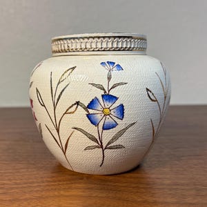 Puede incluir: Un pequeño jarrón redondo de cerámica blanquecina con superficie texturizada. El jarrón está decorado con diseños florales azules y rosas y detalles dorados. El borde tiene un borde decorativo y texturizado.