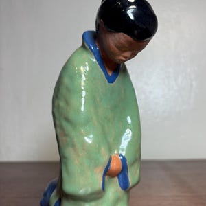 May include: A ceramic figurine of a person kneeling, head bowed. The figure wears a green robe with blue trim and a black head covering. The figurine is on a wooden surface.