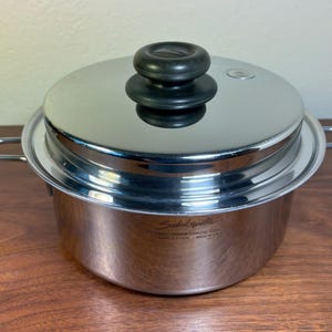 Op de afbeelding: Rvs steel pan met deksel. De pot heeft twee handvatten en een deksel met een zwarte knop. De pot staat op een houten oppervlak. De pot heeft de tekst "Shakespeare" op de zijkant.