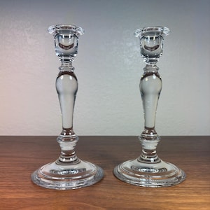 Puede incluir: Dos candelabros de cristal transparente con un diseño clásico. Cada candelabro tiene una base redonda, un tallo delgado con un centro bulboso y una pequeña copa en la parte superior. Se exhiben sobre una superficie de madera.