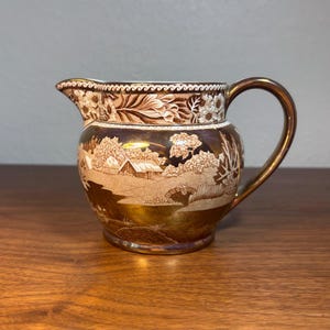 May include: A ceramic creamer jug featuring a metallic copper lustre finish. The body is decorated with a detailed transferware scene of a rural landscape with houses, trees, and a bridge. A decorative border pattern encircles the rim.