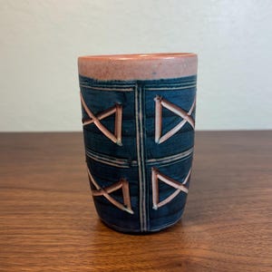 May include: A cylindrical ceramic cup with a dark blue background and a geometric pattern of intersecting lines in a lighter brown color. The rim is a light reddish-brown color.