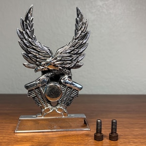 Könnte beinhalten: Silberfarbene Motorradmotor-Ornament mit einem Adler mit ausgebreiteten Flügeln. Der Adler hat detaillierte Federn. Zwei kleine Schrauben befinden sich neben dem Ornament. Die Basis des Ornaments ist rechteckig.