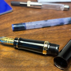 Pu&ograve; includere: Una penna stilografica nera con dettagli dorati, parzialmente smontata su una superficie di legno. Si vede il pennino d'oro, insieme al cappuccio e alla cartuccia d'inchiostro. Un quaderno blu &egrave; sullo sfondo. &Egrave; uno strumento di scrittura.