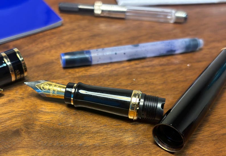 Pu&ograve; includere: Una penna stilografica nera con dettagli dorati, parzialmente smontata su una superficie di legno. Si vede il pennino d'oro, insieme al cappuccio e alla cartuccia d'inchiostro. Un quaderno blu &egrave; sullo sfondo. &Egrave; uno strumento di scrittura.