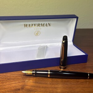 Pu&ograve; includere: Una penna stilografica Waterman nera con dettagli dorati e pennino dorato, accanto alla sua scatola di presentazione blu. La scatola &egrave; aperta, rivelando un interno bianco e il logo Waterman Paris.
