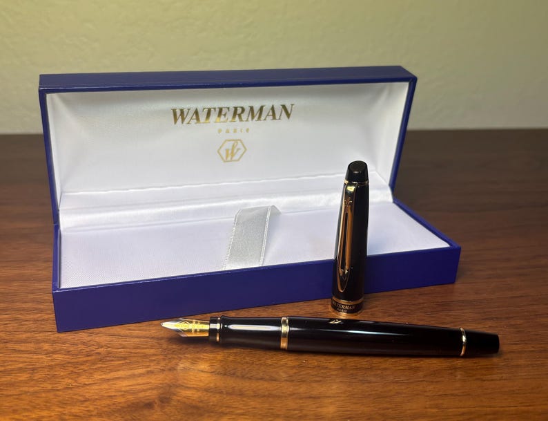 Pu&ograve; includere: Una penna stilografica Waterman nera con dettagli dorati e pennino dorato, accanto alla sua scatola di presentazione blu. La scatola &egrave; aperta, rivelando un interno bianco e il logo Waterman Paris.