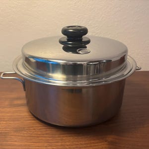 May include: A stainless steel cooking pot with a lid. The pot has two side handles and a lid with a black knob. The pot is sitting on a wooden surface. The pot is designed for cooking.
