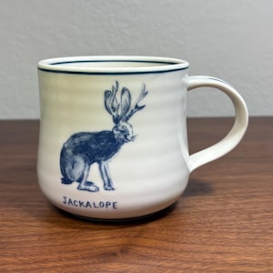 Puede incluir: Taza de cer&aacute;mica blanca con una ilustraci&oacute;n azul de un jackalope y la palabra "JACKALOPE". La taza tiene forma redondeada, un asa y un borde azul. Ideal para bebidas calientes.