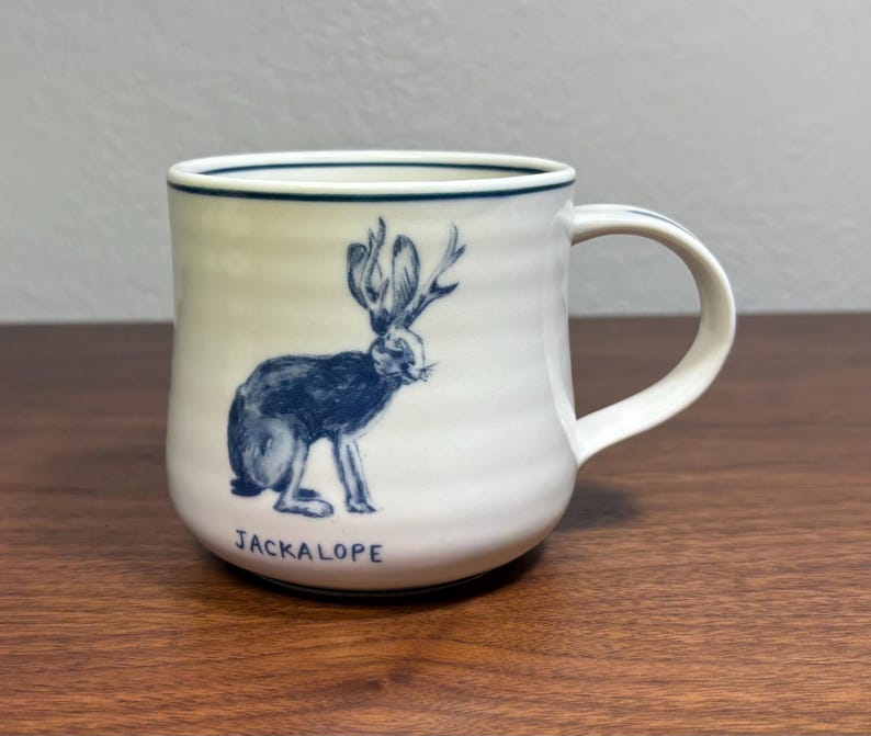 Puede incluir: Taza de cer&aacute;mica blanca con una ilustraci&oacute;n azul de un jackalope y la palabra "JACKALOPE". La taza tiene forma redondeada, un asa y un borde azul. Ideal para bebidas calientes.