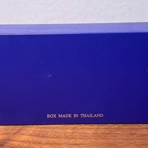 Pu&ograve; includere: Una scatola rettangolare blu cobalto. La scatola ha una superficie liscia e il testo "BOX MADE IN THAILAND" &egrave; stampato in lettere dorate sul fondo. La scatola &egrave; appoggiata su una superficie di legno.