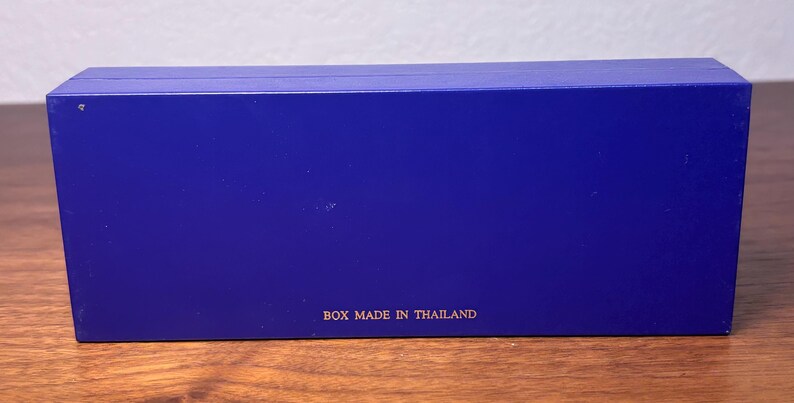 Pu&ograve; includere: Una scatola rettangolare blu cobalto. La scatola ha una superficie liscia e il testo "BOX MADE IN THAILAND" &egrave; stampato in lettere dorate sul fondo. La scatola &egrave; appoggiata su una superficie di legno.
