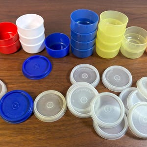 May include: A selection of colourful plastic containers and lids. The containers are red, white, blue, yellow, and peach. The lids are white and blue. Some containers are stacked, others are loose.