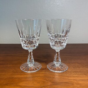 Peut inclure: Deux verres à vin en cristal transparent avec des motifs taillés complexes. Les verres ont un pied et un bol large et légèrement évasé. Le cristal reflète la lumière, créant un effet scintillant. Les verres sont posés sur une surface en bois foncé.