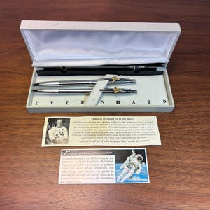 Peut inclure: Un ensemble de stylos Eversharp argentés dans un coffret de présentation blanc avec des emblèmes d'astronaute dorés. Comprend deux cartes informatives avec du texte sur l'alunissage, l'astronaute Gene Cernan et l'histoire de la recharge pressurisée.