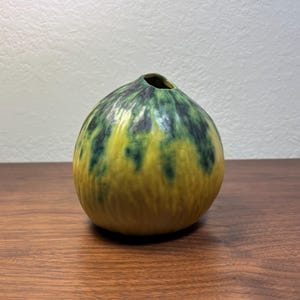 Pode incluir: Um vaso decorativo de cerâmica com formato arredondado e uma pequena abertura no topo. O vaso apresenta uma mistura de cores amarelo, verde e verde escuro, criando uma aparência única e artística. Está sobre uma superfície de madeira.