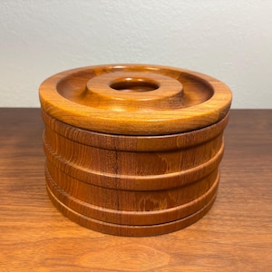 May include: A round, wooden container with a lid. The container is made of light brown wood and has a series of horizontal ridges around its body. The lid has a circular indentation in the center. The container is sitting on a wooden surface.
