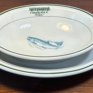 May include: Two white ceramic plates, one stacked inside the other. The top plate has a blue fish illustration and the text "METHAKATIA COMMUNITY HALL" in green. Both plates have a green border.