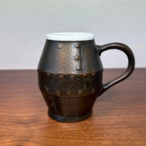 May include: A handcrafted, dark brown ceramic mug with a textured surface and a white rim. The mug features a sturdy handle and is adorned with small, rivet-like details. The mug is designed to hold hot or cold beverages.