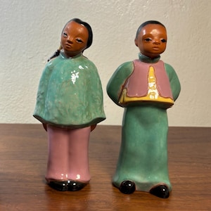 May include: Two ceramic figurines of people in traditional attire. One wears a green cape and pink pants, the other a green tunic with a pink vest. Both have dark hair and black shoes. The figurines are on a wooden surface.