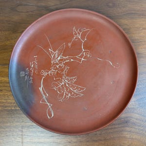 Może przedstawiać: Okrągły, czerwonobrązowy ceramiczny talerz z ręcznie rysowanym kwiatowym wzorem w kolorze białym. Talerz posiada japońską kaligrafię po lewej stronie. Projekt obejmuje kwiat i liście, z delikatnymi liniami i detalami.