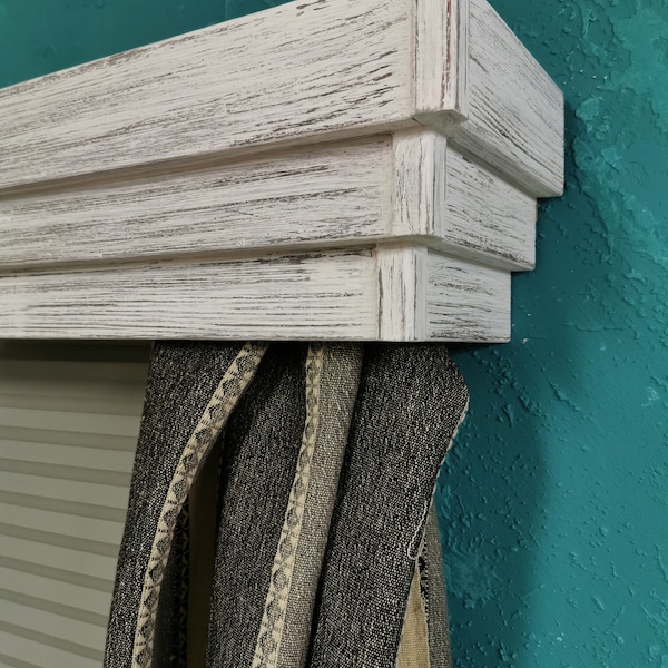 Puede incluir: Cornisa de ventana de madera pintada de blanco con un diseño en capas. La cornisa está montada sobre una ventana con cortinas rayadas en blanco y negro.