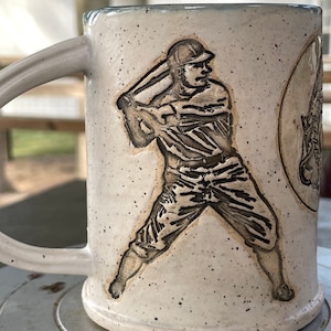 May include: A white ceramic mug with a baseball player design. The player is depicted in a batting stance, holding a bat and wearing a baseball cap. The mug has a speckled glaze and a light blue rim.