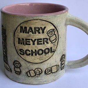 May include: A white ceramic mug with a pink interior and a black inscription that reads "MARY MEYER SCHOOL". The mug is decorated with small black illustrations of children.