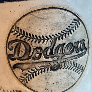 May include: A white ceramic mug with a blue rim and a baseball-shaped design featuring the word "Dodgers" in black lettering.