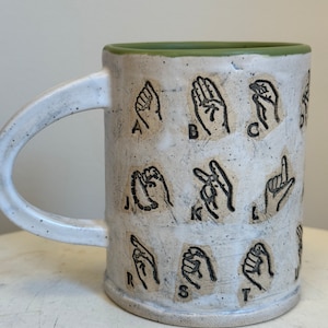 May include: White ceramic mug with a green rim. The mug has black illustrations of the American Sign Language alphabet on the front.