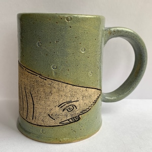 May include: A green ceramic mug with a white shark illustration. The mug has a speckled glaze and a handle.