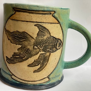 May include: A ceramic mug with a teal glaze and a brown fish design. The fish is inside a round, cream-colored bowl.