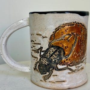 May include: White ceramic mug with a black rim and a brown and black beetle design. The beetle is crawling on a brown, textured surface. The mug has a speckled white glaze.