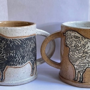 May include: Two ceramic mugs with a speckled glaze. One mug features a black sheep illustration, while the other mug features a white sheep illustration.