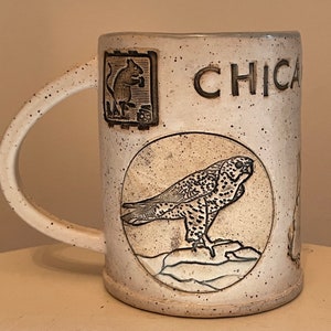 May include: White ceramic mug with a black and white illustration of a bird of prey, a squirrel, and the word "CHICA" on the side.