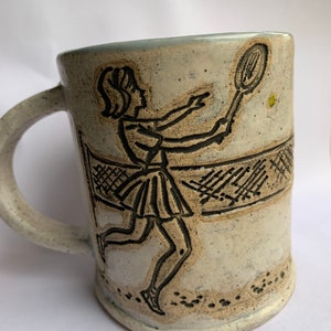 May include: A white ceramic mug with a black silhouette of a tennis player holding a racket and a tennis ball. The mug has a speckled glaze and a brown handle.
