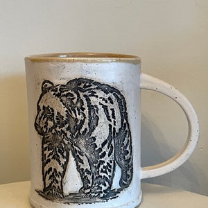 May include: White ceramic mug with a black and white illustration of a bear walking. The mug has a brown rim.