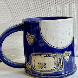 May include: A blue ceramic mug with a white and speckled design. The mug features a white circle with a black outline and a jar with fireflies inside. The jar is surrounded by fireflies.
