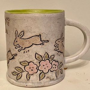 May include: White ceramic mug with a green rim and a hand-painted design of brown rabbits running through a field of pink flowers and green leaves.