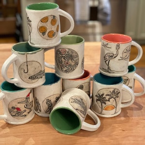 May include: A stack of ten white ceramic mugs with different illustrations. The mugs have green, blue, and red rims. The illustrations include a fish on a hook, a flamingo, palm trees, a pelican, and a group of people.