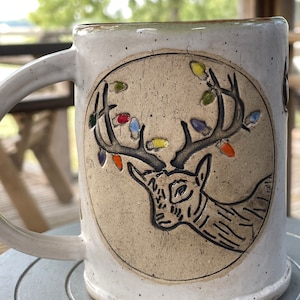 May include: White ceramic mug with a brown deer head design and colorful lights on the antlers. The mug has a round handle.