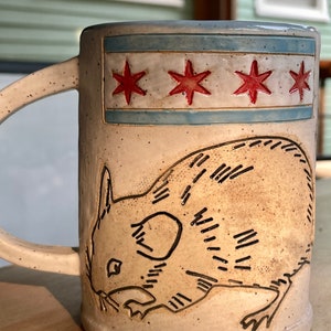 May include: A white ceramic mug with a blue and red Chicago flag design and a black line drawing of a rat on the front.