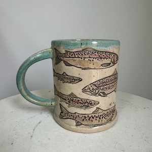 May include: A ceramic mug with a teal rim and handle features a design of five brown trout fish on a cream background.