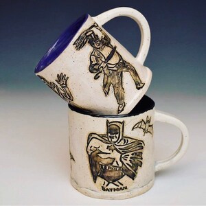 May include: Two ceramic mugs with black and white illustrations. The top mug has a blue rim and features the Penguin from Batman. The bottom mug features Batman and a bat. The bottom mug has the word "BATMAN" written on the bottom.