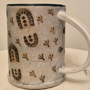 May include: White ceramic mug with a black rim and a pattern of brown footprints and small branches.