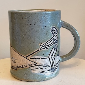 May include: A green ceramic mug with a white and black illustration of a person water skiing. The mug has a speckled glaze and a white interior.