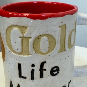 May include: White ceramic mug with a red rim. The mug has the words "Gold Life Master" written on it in black and yellow lettering.